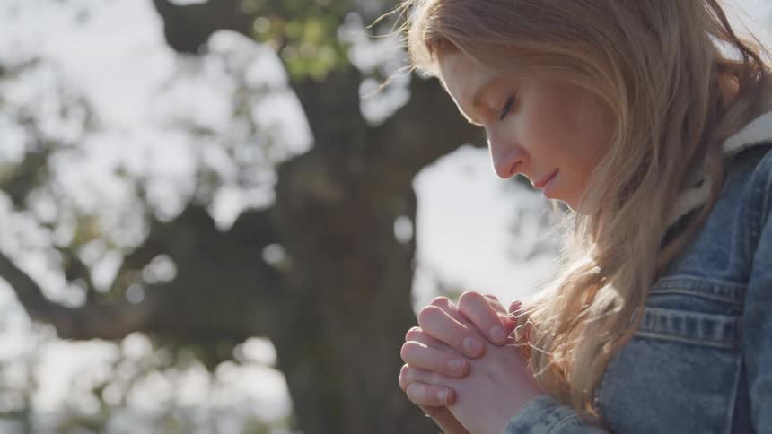 A woman with long light brown hair clasps her hands together in contemplation her head bowed in a moment of quiet reflection with a blurred natural background