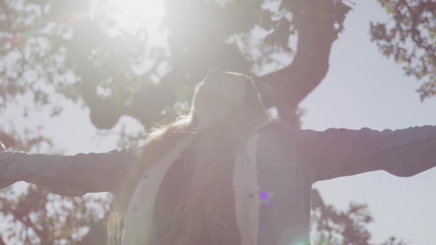 A woman with long light brown hair stands with her arms outstretched facing the bright sunlight filtering through the trees above her