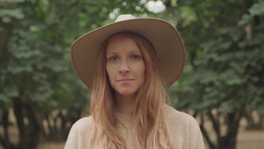 A woman with long reddish brown hair wearing a wide brimmed hat looks directly at the camera with a gentle expression against a blurred background of trees
