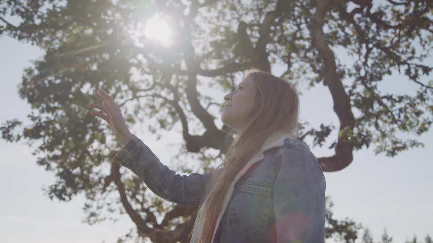 Blonde woman standing outdoors near a tree smiles while raising a hand towards the sky