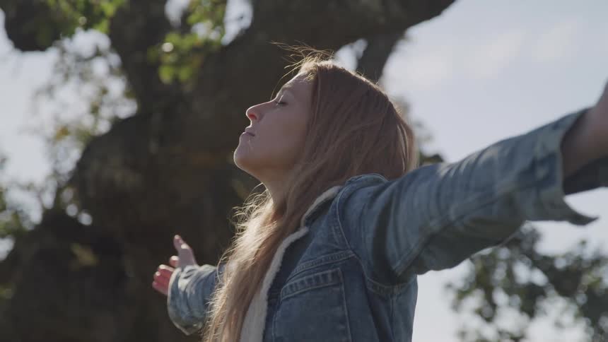 Woman with long hair standing with arms wide open and eyes closed in front of large tree