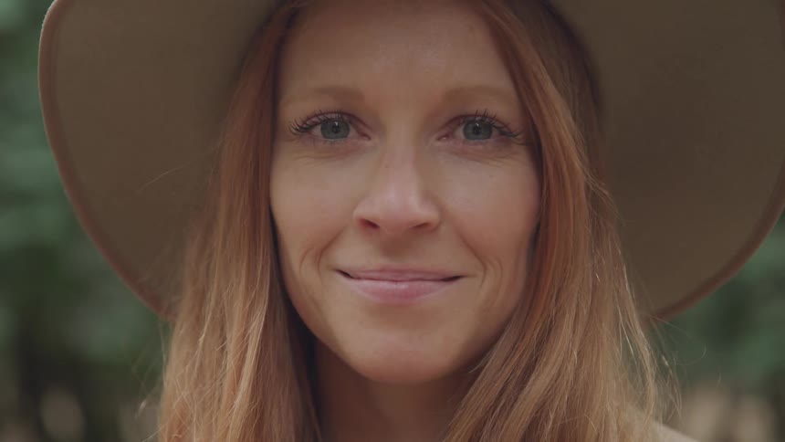 A close up shot captures the face of a woman with long reddish brown hair fair skin and blue eyes wearing a wide brimmed hat and smiling gently at the camera