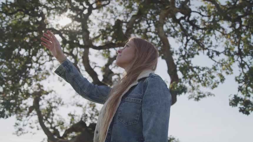 Woman lifts her head and reaches one hand toward the sky with a slight smile on her face