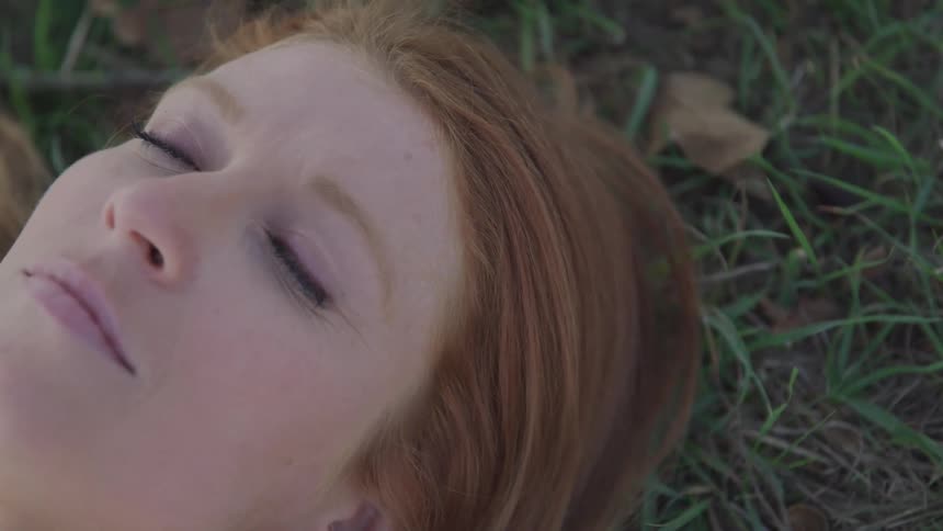 A woman with reddish brown hair lying on the grass with her eyes closed