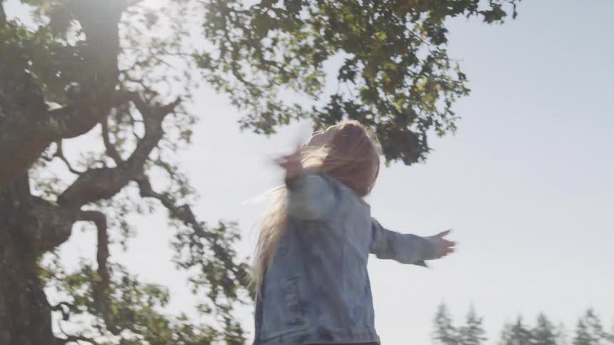 A woman with long light brown hair is captured mid turn with her arms outstretched and her gaze directed upwards towards the sky against a backdrop of tree branches
