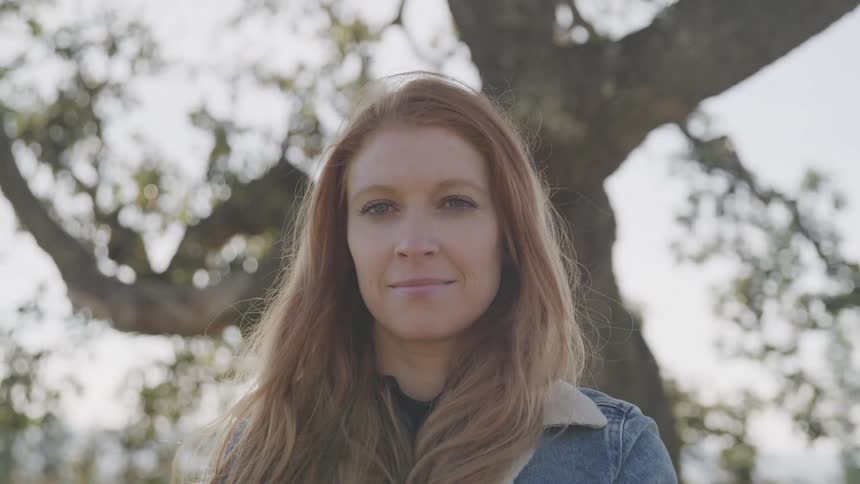 A young woman with long reddish blonde hair and a denim jacket smiling slightly at the camera with a blurred tree branch in the background