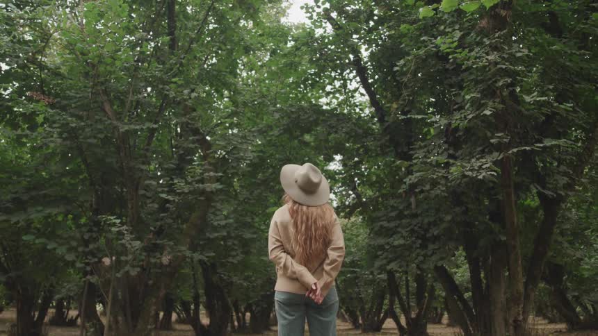 A woman with long light brown hair wearing a hat and light colored clothing stands with her back to the camera hands clasped behind her facing a dense hazelnut orchard