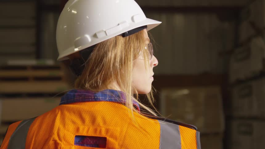 Woman wearing a hardhat and a safety vest walks through a warehouse