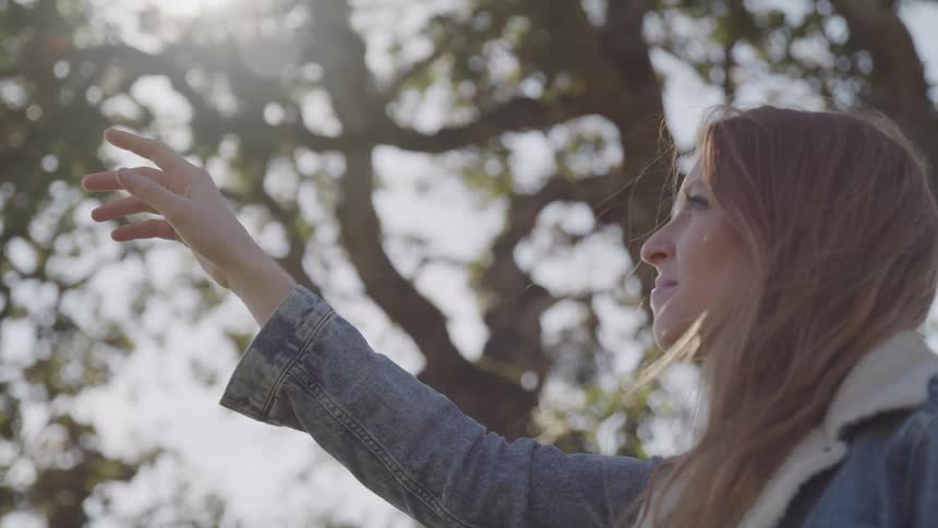 A woman with long light brown hair wearing a denim jacket reaches her arm upwards towards the sunlight filtering through the trees