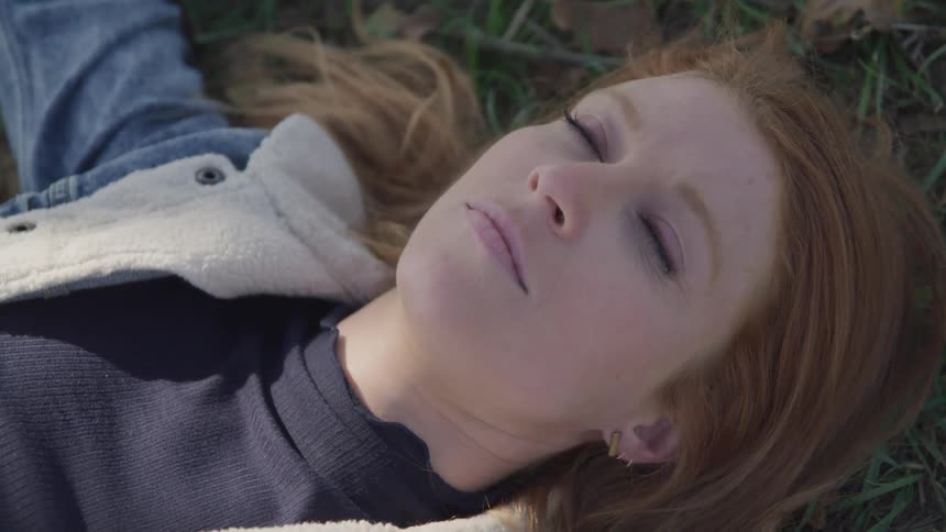A woman with light brown hair and fair skin lies on the grass gazing upwards with her eyes open wearing a denim jacket and a dark top