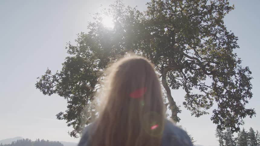 A woman with long light brown hair stands with her back to the camera facing a large tree silhouetted against a bright sunlit sky