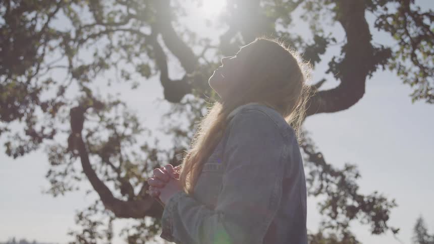 A woman with long light brown hair stands with her hands clasped looking up towards the sunlight filtering through the branches of a large tree