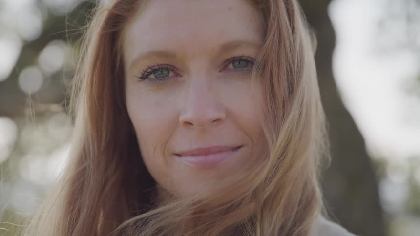 A woman with long light brown hair fair skin and blue eyes with a gentle smile and a blurred natural background