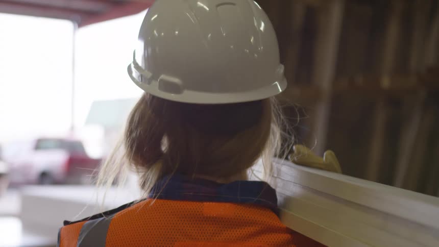 A worker wearing a hardhat is carrying some 1by4 studs on her shoulder in a warehouse