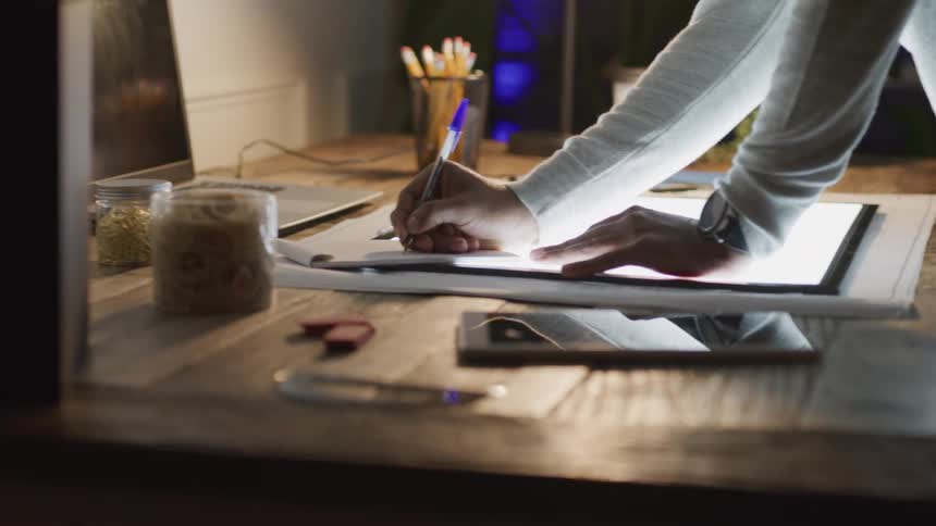 Person writing on paper covering a very large brightly lit tablet on the table