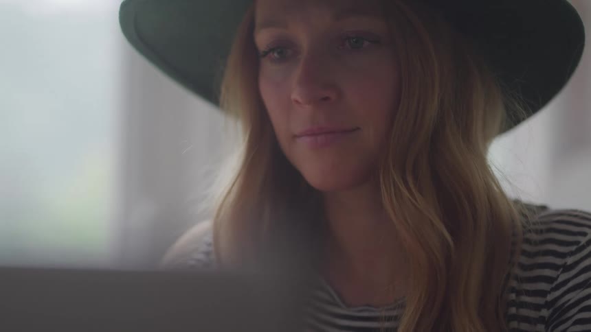 A woman with long light brown hair and a dark hat using a laptop with a thoughtful expression with a blurred background creating a sense of intimacy