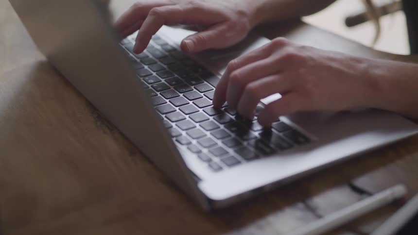 A closeup captures a person typing on a macbook air at a wooden table as the frame pans to reveal an apple pencil and part of an ipad