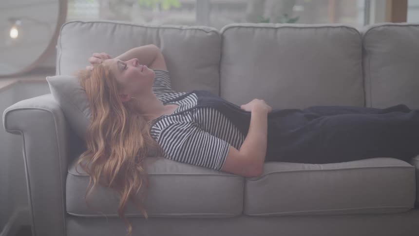 A woman with long reddishbrown hair lies on a gray couch with her eyes closed and her arm resting behind her head appearing contemplative