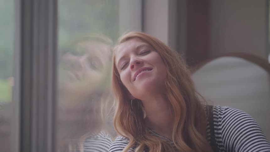 A young woman with long reddish blonde hair leans against a window looking up thoughtfully before closing her eyes with a peaceful smile