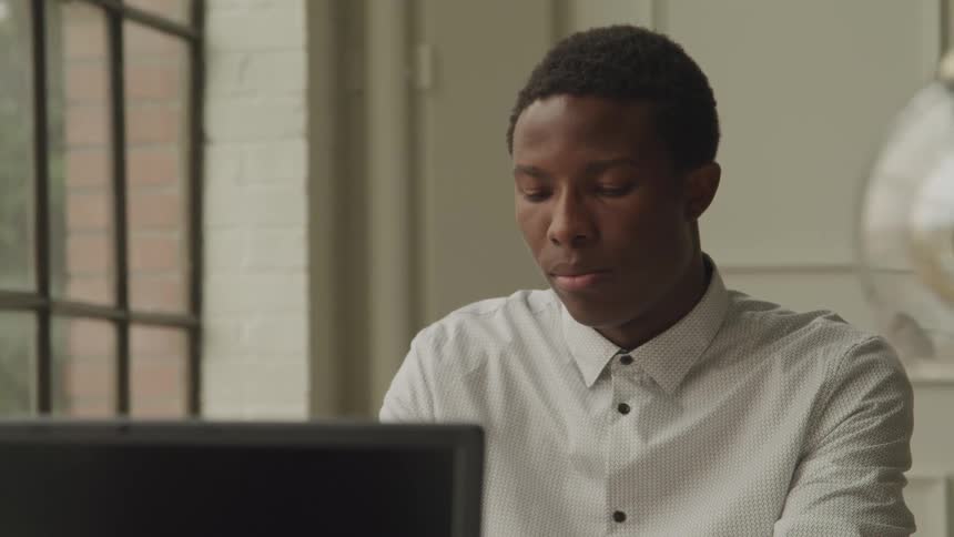 A male is typing on his laptop and sitting next to a window