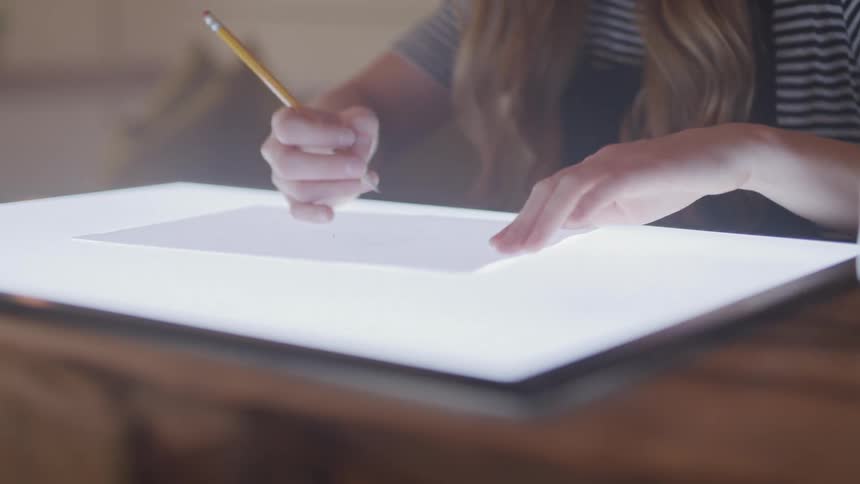 A girl sits at a desk tracing paper on a light board