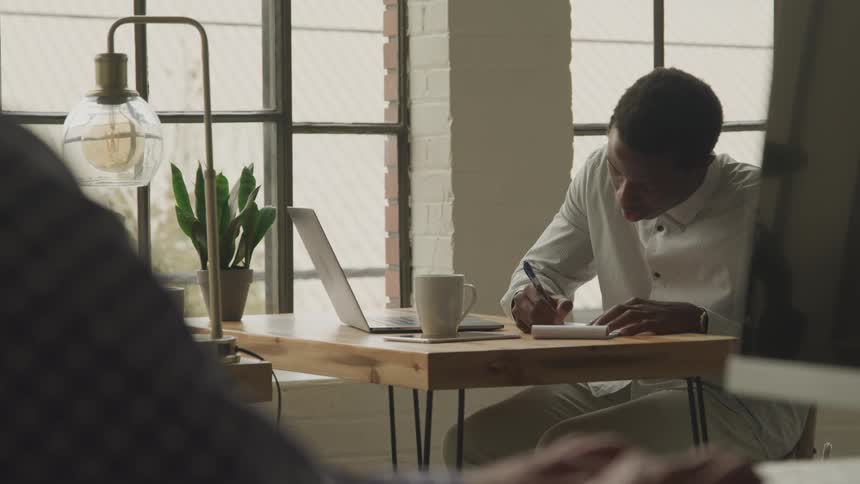 A man types on his laptop and writes notes in a pad