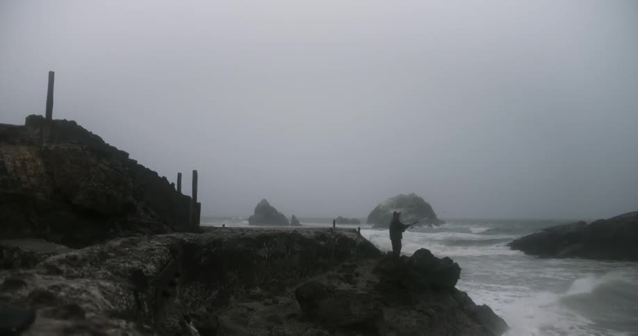 A man stands on a rocky coastline while fishing on a foggy morning