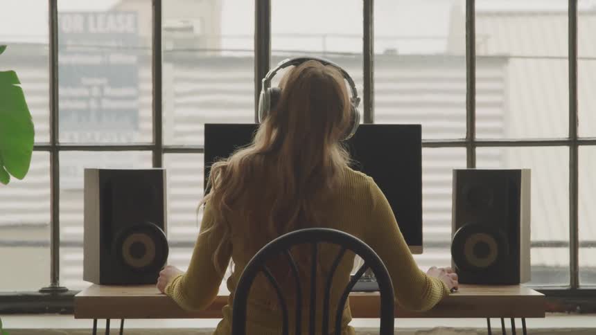 Blonde woman puts on headphones and dances in her seat in front of a computer