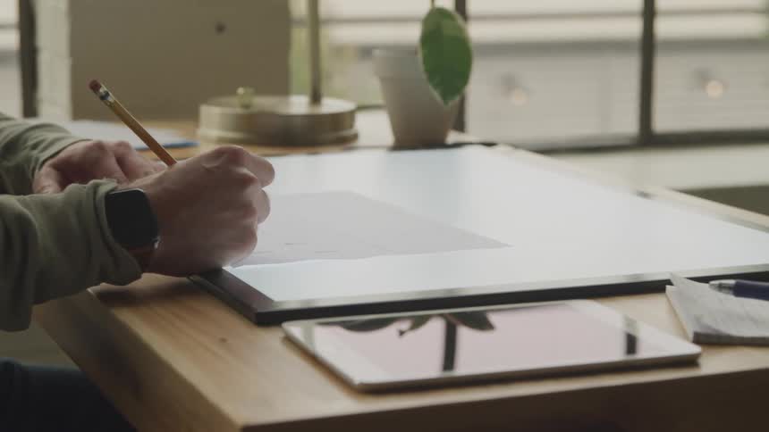 Person draws with a pencil while sitting at a desk near a tablet