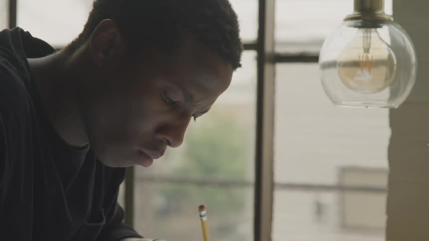 The black man is looking down and writing something with the pencil he is holding