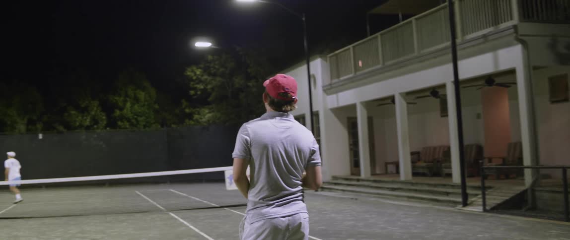 A man stands by a tennis court waiting for the opposite player at night