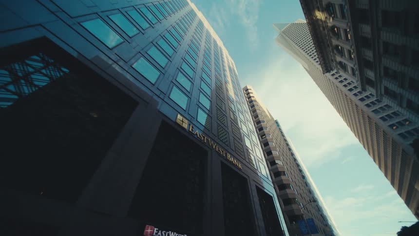 A modern skyscraper with a dark facade and reflective blue windows angled upwards towards a blue sky with faint clouds with the corner of another taller building visible on the right