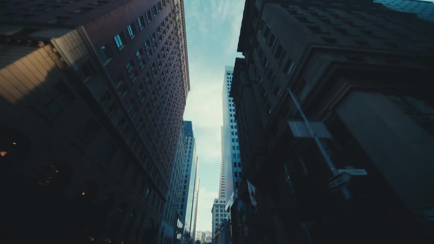 A narrow city street framed by tall dark buildings revealing a sliver of bright sky above