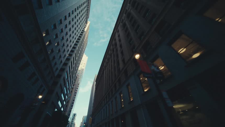 A low angle view shows the towering height of buildings lining a city street with a narrow strip of sky visible between them
