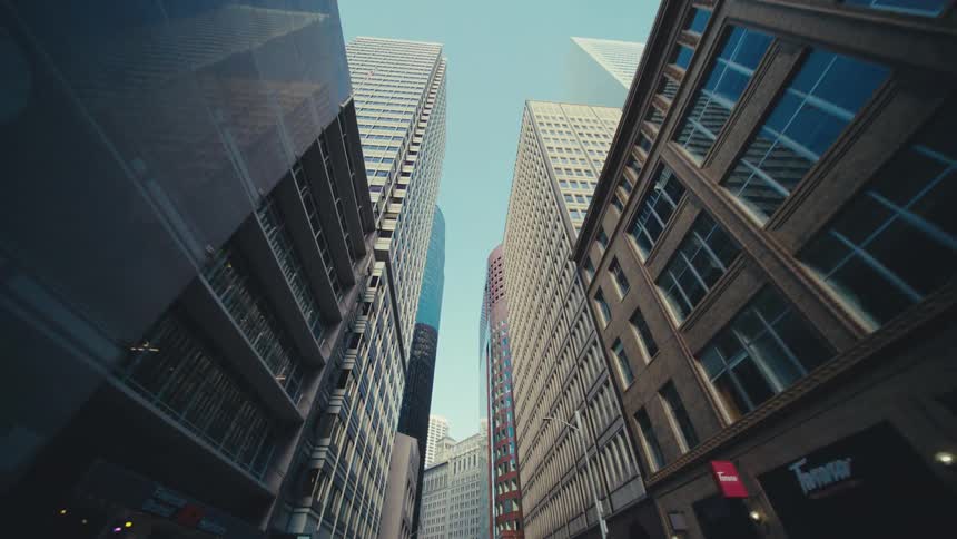 Tall buildings framing a narrow street revealing a sliver of blue sky above with the buildings displaying a mix of architectural styles and colors including glass and brick elements