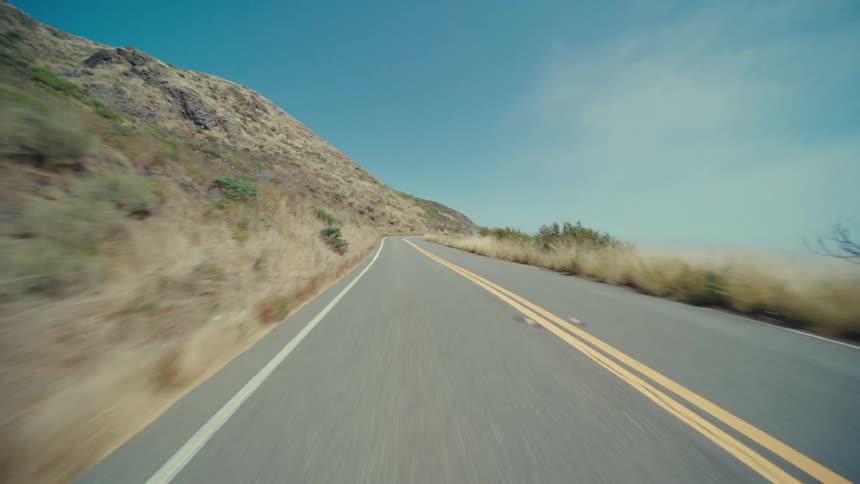 A straight road with double yellow lines stretches into the distance ascending a hill with a dry grassy slope on one side and a hint of the ocean visible on the other under a clear blue sky