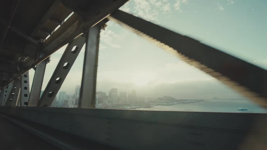 A coastal metropolis comes into view through steel beams as seen from a moving vehicle traveling the lower level of a bridge
