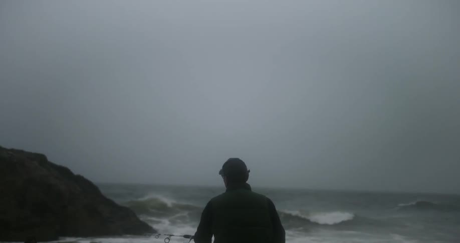 A man fishing on the coast next to a large boulder