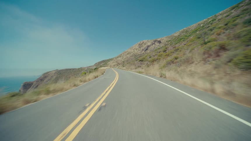 A straight empty road stretches into the distance under a clear blue sky flanked by a grassy slope on one side