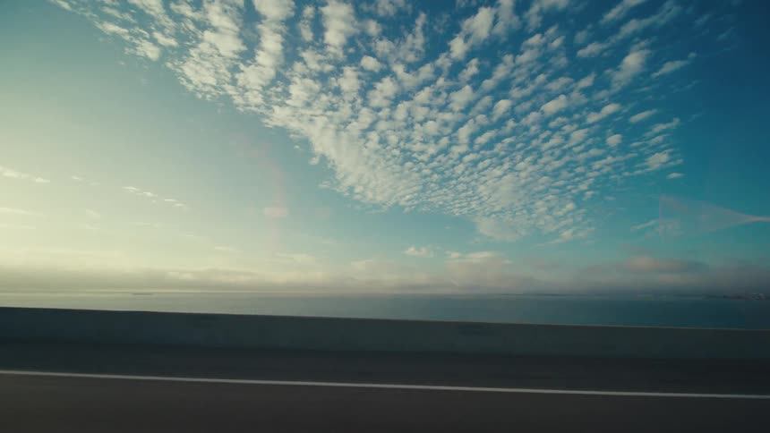 View from a moving vehicle on a bridge showing expansive ocean and dramatic cloud formations with the bridges concrete barrier in the foreground