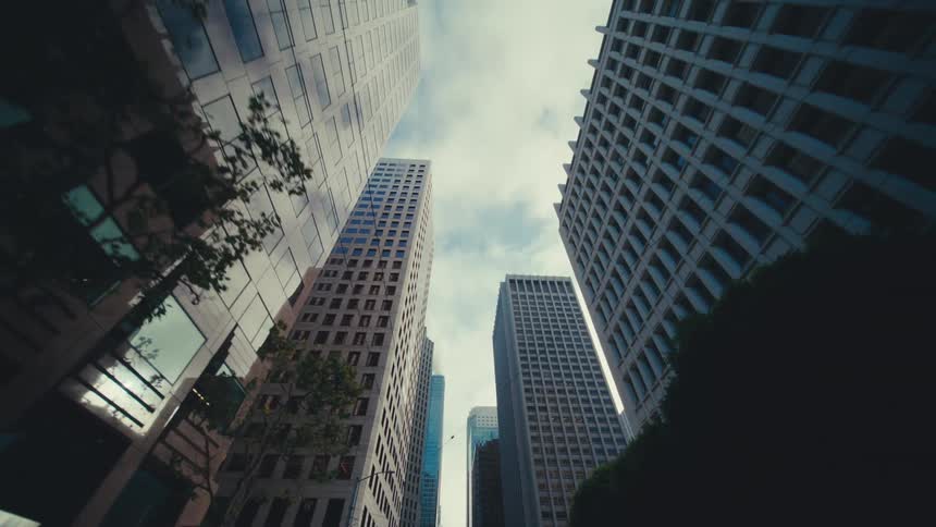 A streetlevel upward view glides past towering skyscrapers and occasional trees with overhead power lines and glass windows reflecting the cloudy sky