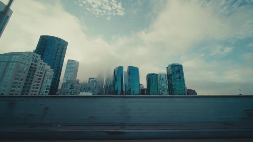 A cityscape with tall buildings is seen from a moving vehicle with a cloudy sky and a concrete barrier in the foreground