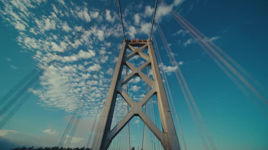 A suspension bridge travels under a bright blue partially cloudy sky
