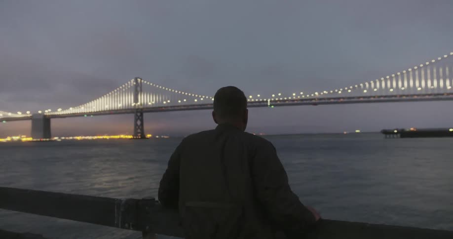 A man standing on a pier looking out of the water with a bridge and it is lit up
