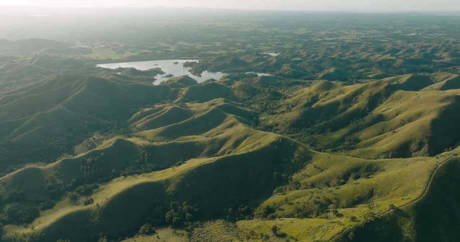 An aerial view of expansive rolling hills stretches toward a distant lake revealing a sunlit landscape of green terrain and subtle elevation shifts