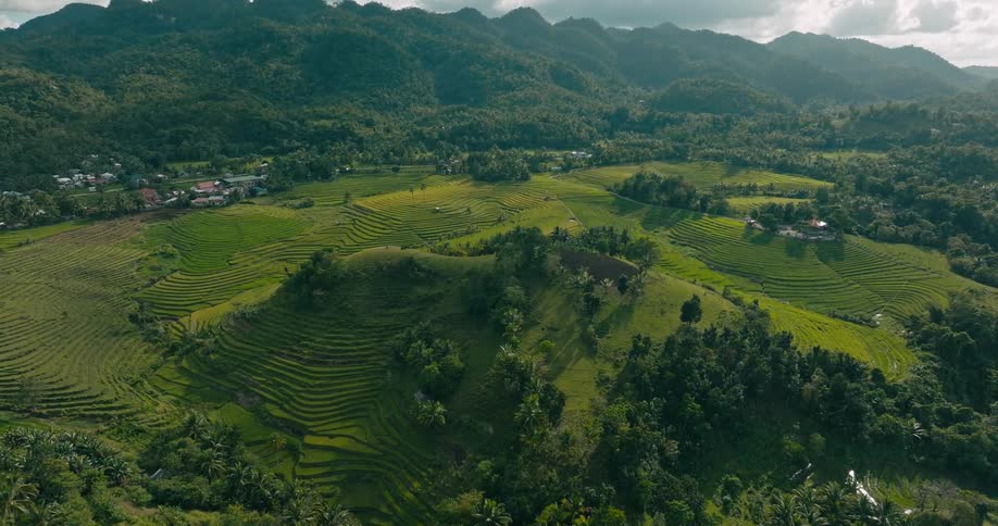 An aerial view reveals patterned farm fields flowing into a remote rural town surrounded by lush hills and terraced green landscapes