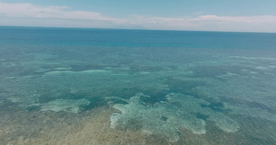 An aerial view captures the shallow clear waters of the open ocean revealing coral formations just beneath the surface