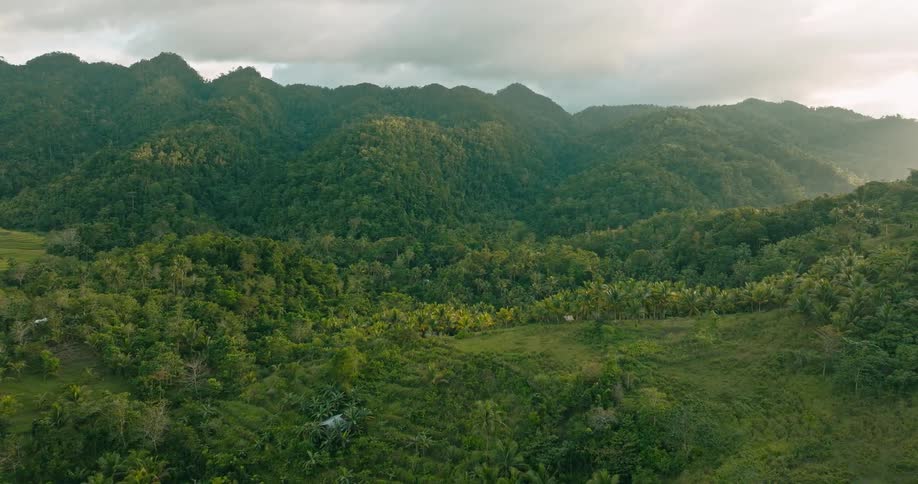 An aerial view rises and glides forward over a lush tropical mountain range covered in dense forest under a cloudy sky