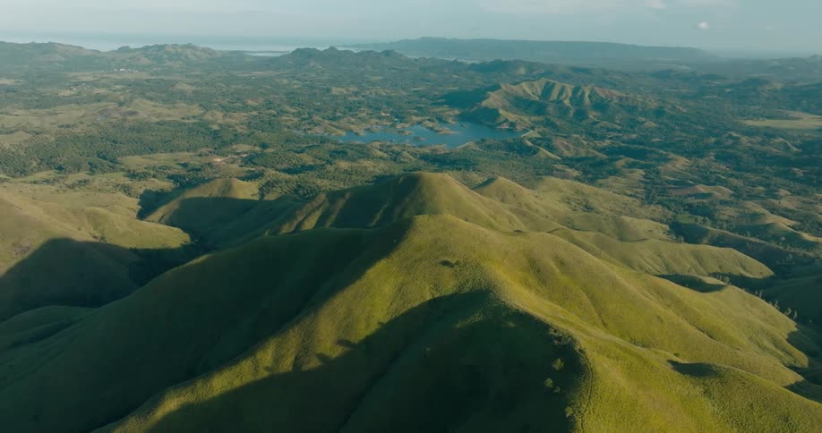 An aerial view sweeps over lush green mountains revealing a distant lake nestled between the ridges