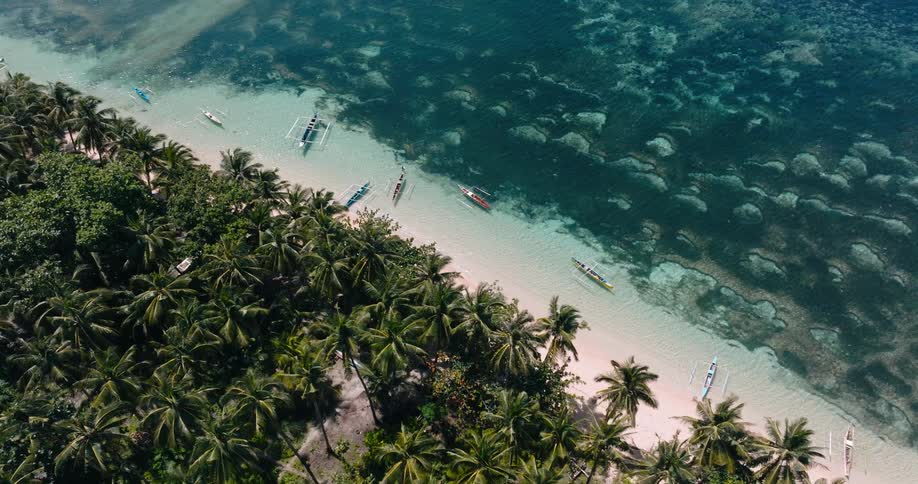 The sweeping aerial view captures the coastline of a tropical island where palm trees line a pristine white beach scattered with boats as people walk along the shore and another boat drifts into frame over crystalclear water